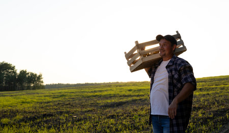 The farmer looks in surprise towards the empty space for insertion while standing at sunset in a field with a box of potatoes. The concept of agriculture, harvesting. A man rejoices in the harvest.の写真素材