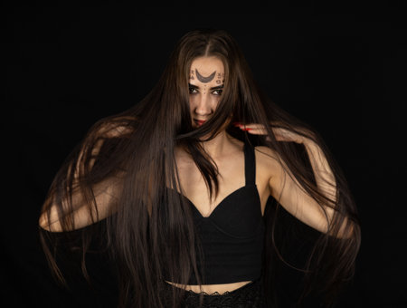For halloween, a female witch stands in the dark with tousled hair falling over her face. Portrait of a Vedbma with gothic symbols on her face and long black hair. Witch concept for halloween.の写真素材