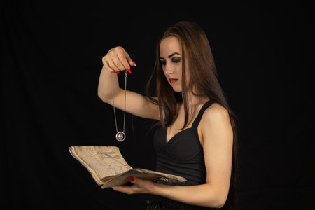 On Halloween, the witch performs a ritual with a book and a talisman. A beautiful female witch reads fortunes from an ancient book, holding a talisman in her hand. Halloween divination concept.の写真素材