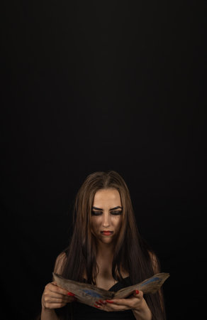 vertically. On the night of halloween, the witch carefully reads a book with spells while standing on a black background. A beautiful witch woman holds an old book in front of her and reads.の写真素材