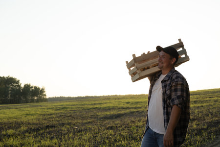 A cute smiling farmer carries a box of potatoes along the edge of the field against the background of the sunset. Copy space. A happy farmer has finished harvesting and is smiling at sunset.の写真素材