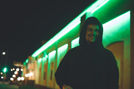 On the street of a city at night, a handsome man laughs. A man in a hood smiles sweetly at night in the neon light of the city. City nightlife concept.の写真素材