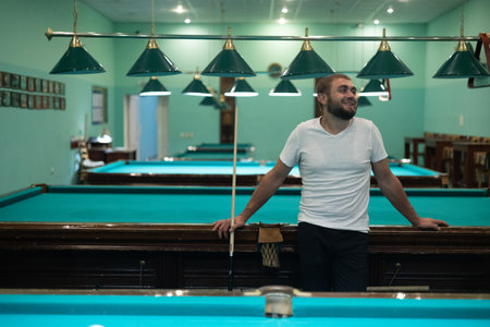 A happy handsome man stands in the billiard room, leaning on the table and chatting with friends. The man is having a blast having fun with his friends and playing billiards.の写真素材