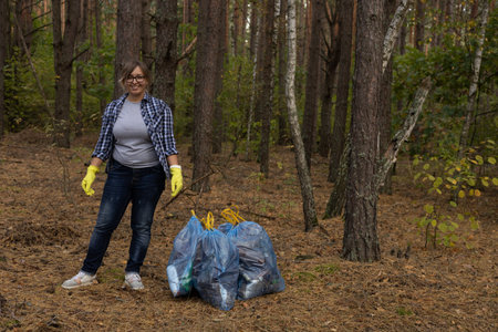 A female volunteer stands in the forest with garbage collected in a bag and looks at the camera smiling. Copy space. A woman in a plaid shirt, an eco activist, collected plastic waste in the forest.の写真素材