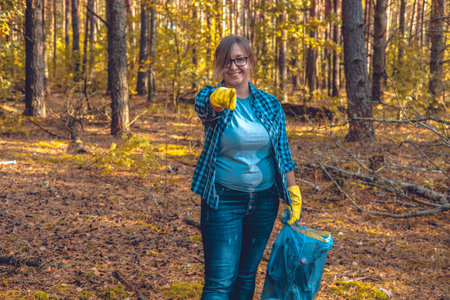 A female volunteer smiles while standing in the forest with a garbage bag in her hand and points her finger at the camera. The woman urges that you too can help save nature from garbage.の写真素材