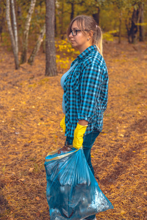 A tired female volunteer after cleaning up garbage in the forest stands with a garbage bag in her hands and looks sadly to the side. A woman worries about food and its pollution by garbage waste.の写真素材