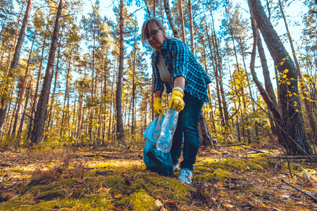 A woman with a garbage bag collects plastic garbage in the autumn forest. Saving the planet from garbage. A woman wearing protective gloves fights to save the planet from garbage.の写真素材