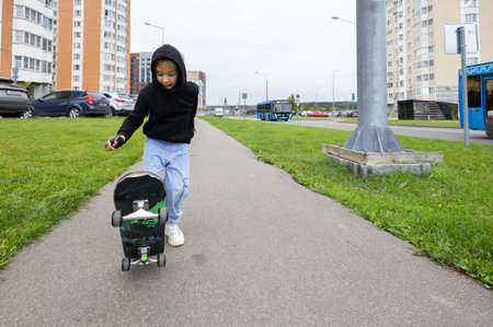 A beautiful child rides a skateboard along the city sidewalk. A child in a hood on a skateboard walks in the city during the day. Childhood of children in the city. City life.の写真素材