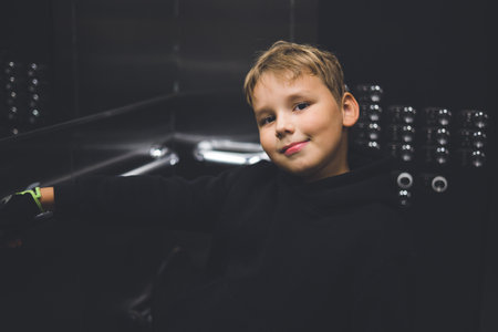 A cute boy stands in an elevator and smiles at the camera near the mirror. A child in a sweatshirt rides in an elevator. Portrait. A beautiful child rides an elevator and smiles at the camera.の写真素材