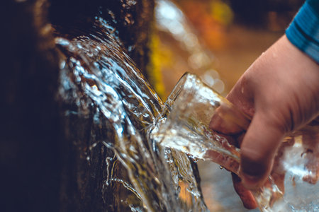 A hand brings a transparent bottle to a spring of drinking water. A man collects mineral water from a spring in the autumn forest. The traveler stopped at a spring to drink and collect clean water.の写真素材
