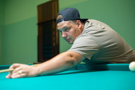 A serious man holding a cue in his hands aims at a billiard ball. A man plays Russian billiards with his friends while having fun. A man is intently aiming at a ball on a billiard table.の写真素材