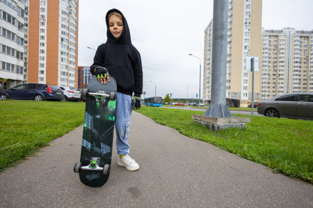 Copy space. On a city street, a stylish boy stands with a skateboard. A boy holds a skateboard with his hand during the day on the street among houses and cars. A child rides a skateboard.の写真素材