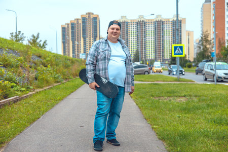 A handsome man stands with a skateboard in his hand and smiles at the camera on a city street. A man in a plaid shirt stopped to skateboard. Concept of a man with a skateboard.の写真素材