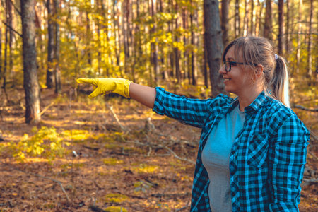 A woman smiles with garbage collected in the forest and points her gloved finger towards the empty space for an inscription. Copy space. A woman takes the initiative to save the forest from garbage.の写真素材
