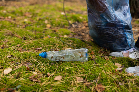 A gloved hand picks up a plastic bottle from the ground and places it in a trash bag. A volunteer man removes trash scattered in the forest. Eco, saving nature, eco activism.の写真素材