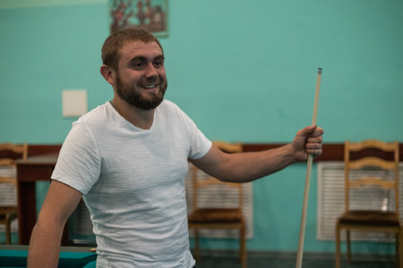 Leaning on the billiard table, the man laughs cheerfully, holding a cue in his hands. A man relaxes and laughs while playing billiards with his friends. Concept of a fun game of billiards.の写真素材