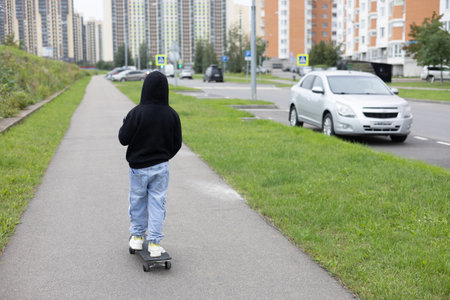 View from the back. A child pushes his foot off the sidewalk and rides a skateboard. Lifestyle. A boy in a hooded sweatshirt rides a skateboard along the city streets.の写真素材