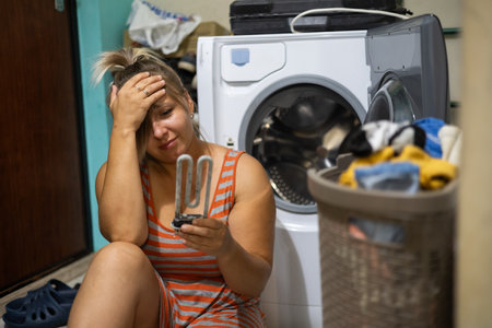 An upset woman holding her head sits near a broken washing machine with a heating element in her hand. A woman with a basket of dirty laundry grieves near the washing machine at home.の写真素材