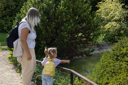 Mother and daughter stand on the shore of a decorative pond and admire the swimming fish. In a beautiful park, mother and daughter are relaxing near a fish pond. Family on vacation concept.の写真素材