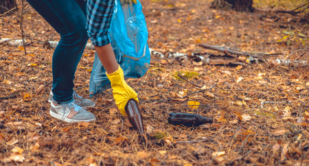 A woman in the forest bends down and picks up glass bottles from the ground and puts them in trash bags. Eco activism, nature protection, ecology. A woman volunteer saves the forest from garbage.の写真素材