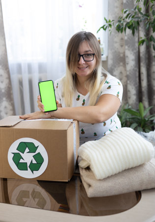 Vertical. A woman shows a green phone screen over a box of clothes for donation. Advertising for...