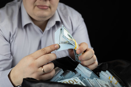 On a black background, a man flips through bills in a stack of dollars above a bag with money. A man...