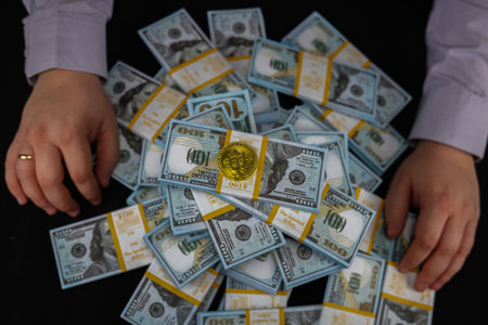 A trader holds his hands on either side of a stack of dollar bills with a bitcoin coin on top. The...