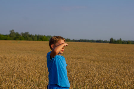 In a wheat field on a summer day, a child looks into the distance, covering his eyes with his hand. Shielding his eyes from the bright summer sun, the child looks at the wheat field.の写真素材