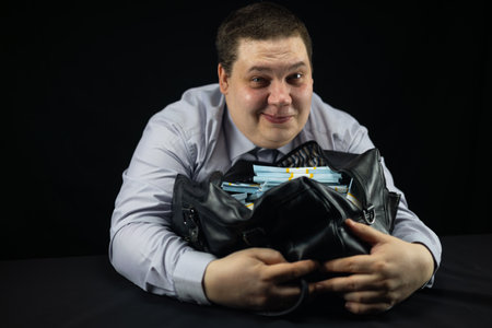 A male official hugged a bag full of dollars and looked happily at the camera on a black background. A joyful official with a black bag full of stacks of hundred dollar bills.の写真素材