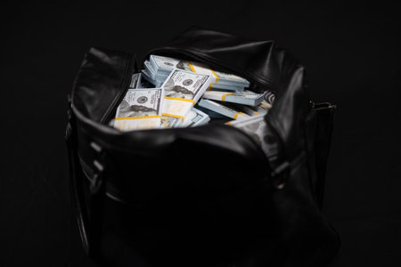 An open bag with a lot of dollars stands isolated on a black background. Through the unzipped zipper of the bag, one can see packed wads of hundred dollar bills. Bag full of money.の写真素材