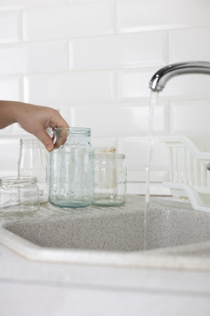 Vertical. Glass sorting. In the kitchen sink, a woman under the tap washes a glass jar to be sent...