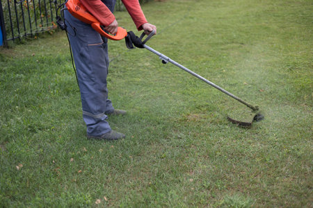 A man mows the grass in front of his house with a trimmer. A man uses an electric trimmer to cut...