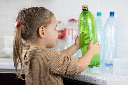 A child enthusiastically helps sort plastic bottles in the kitchen for recycling. A cute little girl takes a plastic bottle from the table to take it to the recycling box.の写真素材