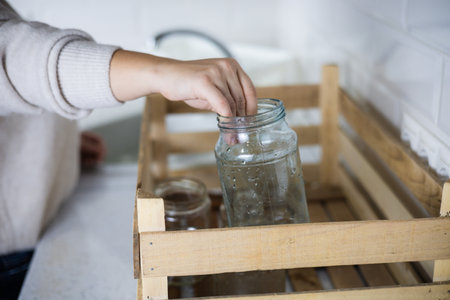 Sorting glass for recycling. A woman in the kitchen carefully places a glass jar into a recycling bin. A woman puts glassware into a recycling bin. Glass sorting concept.の写真素材