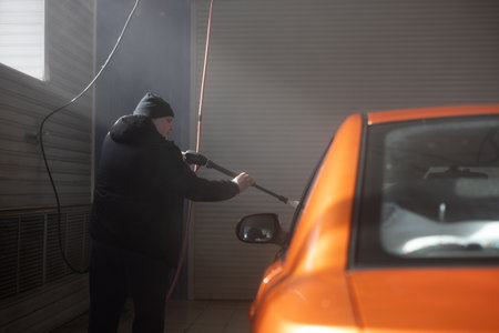 At a car wash, a man washes off dirt and dust from his car. A man walks around a car and washes an orange car with water under pressure.の写真素材