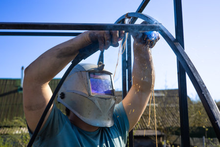 A man welds metal in the garden wearing a protective mask in summer. A man works in his garden...