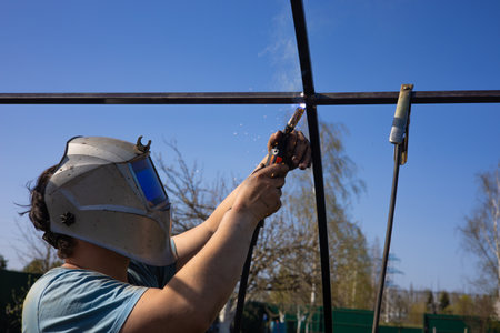 While welding a metal frame for a greenhouse, a man works in a protective mask to protect his eyes...