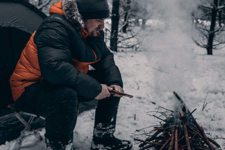 A male tourist in a warm jacket lights a fire near a tent in a snowy forest, amidst a serene winter landscape. Smoke slowly rises into the cold air. A man warms himself by a fire in winter.の写真素材