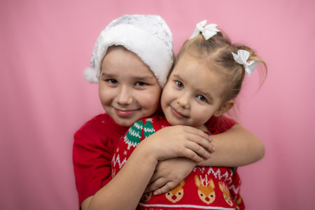 A child in a Christmas hat hugs his sister in a festive sweater on a pink background, radiating happiness and joy. Children rejoice in the coming holidays of Christmas and New Year.の写真素材