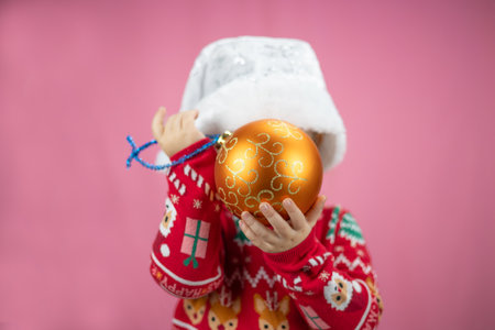 Little child covers face with golden Christmas ball on pink background. Cute baby is wearing festive red and white Christmas sweater and New Year hat. Concept child, christmas, new year.の写真素材