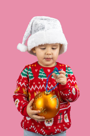 A happy child in a Christmas sweater and Santa hat holds a golden ornament on a pink background. The child conveys the atmosphere of the Christmas and New Year holiday.の写真素材