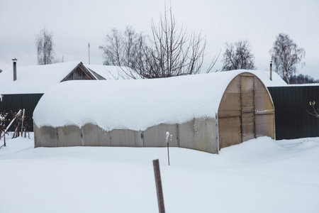 An old greenhouse stands in the backyard of a country house, covered with a thick layer of snow in a cold winter. Beautiful gentle winter in the garden with a covered greenhouse.の写真素材