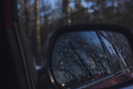 Winter forest in car mirror. Close-up of car rear view mirror reflecting bare trees and soft light of serene winter forest capturing beauty of nature during cold months.の写真素材