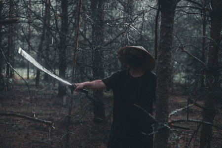 A man trains with a katana. A swordsman wearing a conical Asian hat holds a katana in the forest while it rains. The man practices his sword technique in a dark atmospheric environment.の写真素材