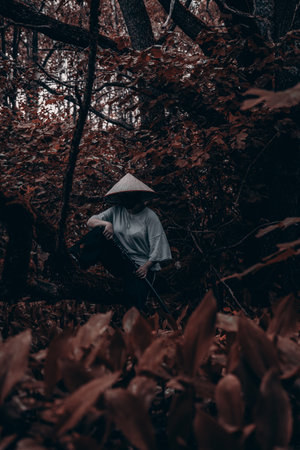 A samurai warrior in a straw hat stands in a dark forest holding a katana sword. A female warrior is...