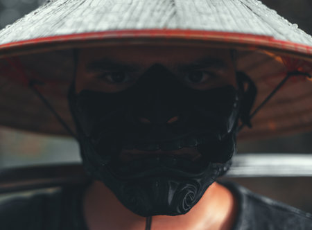 The man in the straw hat and black samurai mask exudes strength and mystery, embodying the essence of a warrior. The dark shadow adds danger, enhancing the fearless presence of the samurai.の写真素材