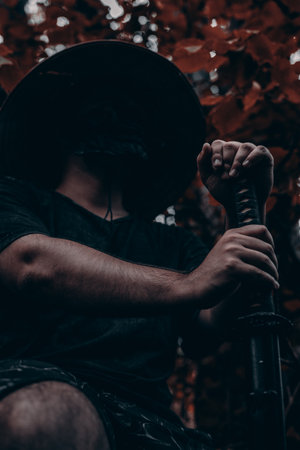 A samurai warrior in a straw hat and mask sits in the forest holding a katana. The warrior with the katana is illuminated by sunlight shining through the foliage.の写真素材