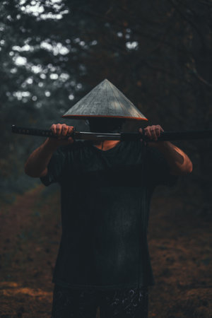A warrior wearing a straw hat and a black mask stands in a fighting pose holding a katana with both hands The warrior with the katana is surrounded by trees and the atmosphere is dark and mysterious.の写真素材