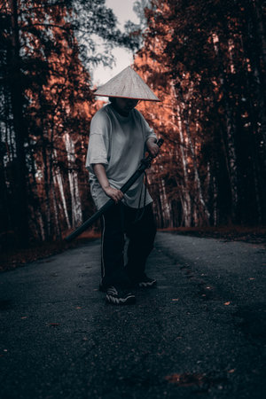 A warrior stands on a path in the forest, holding a katana in both hands. He wears a straw hat and dark clothes.の写真素材