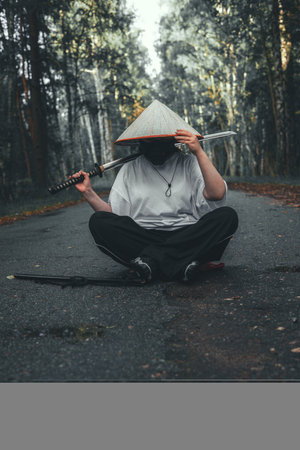 Female warrior with katana. Cosplayer in Japanese style costume sits cross-legged on forest road holding katana with serious expression, creating mysterious and adventurous atmosphere.の写真素材